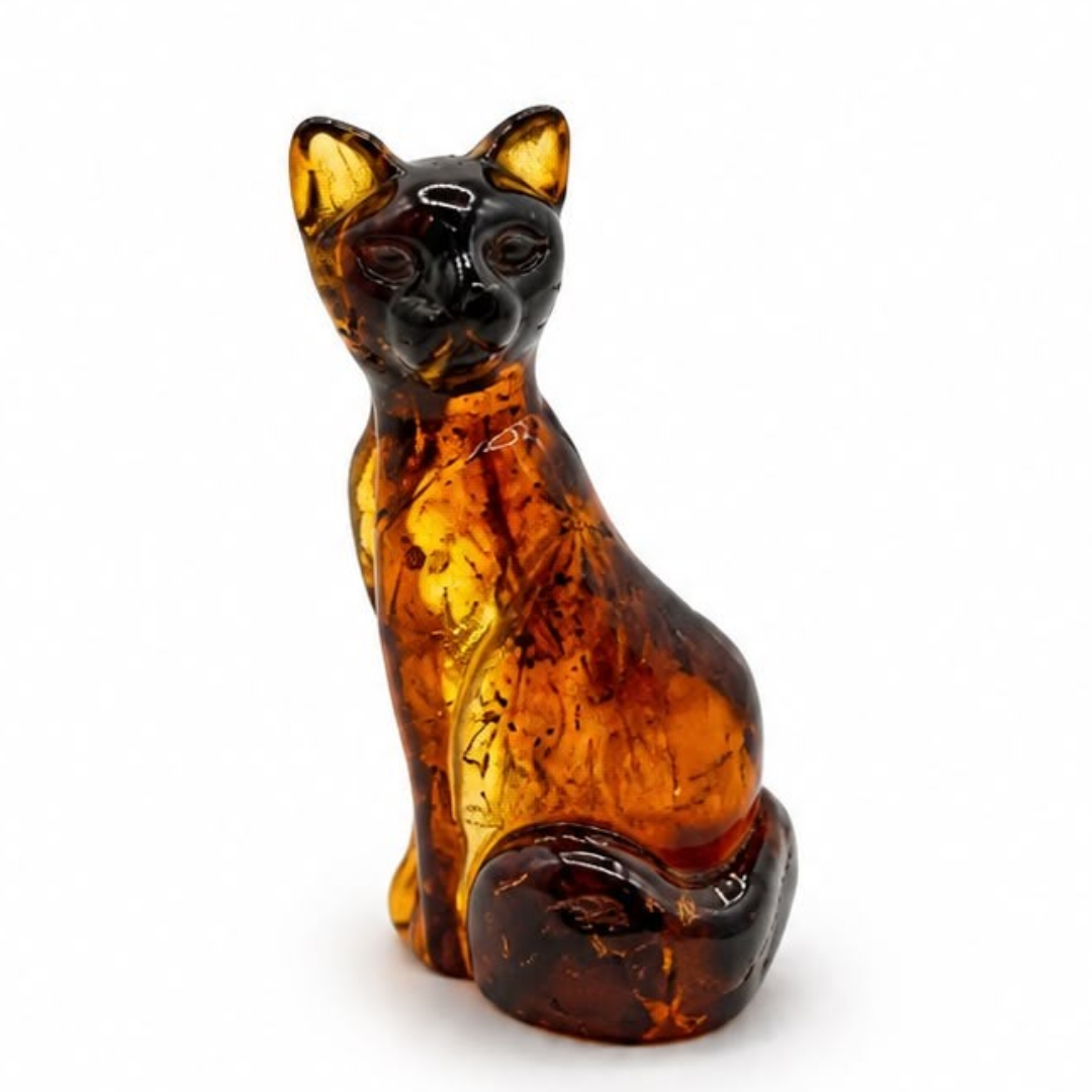 A small amber cat figurine, shown sitting down, made of translucent amber glass with dark brown and golden-yellow highlights, set against a plain white background.